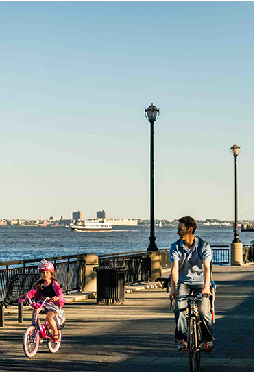 Father and daughter riding bikes along Hudson river waterfront with ferry ships on the water.