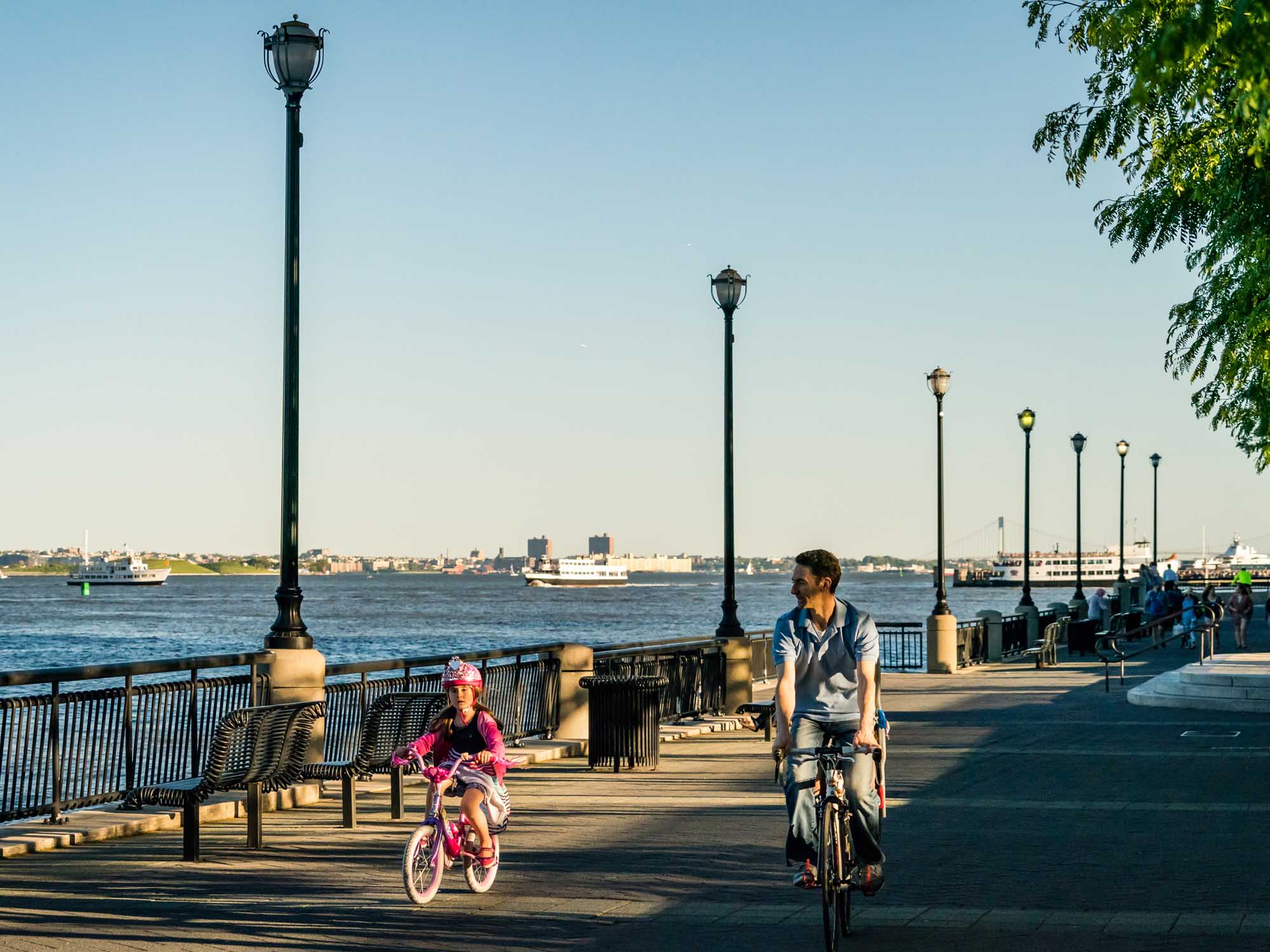 Father and daughter riding bikes along Hudson river waterfront with ferry ships on the water.