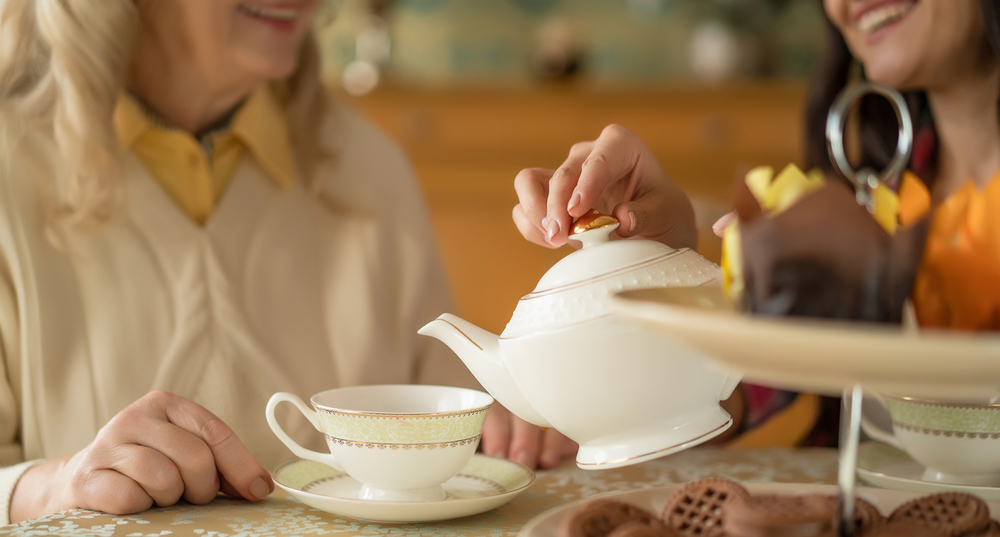 Hands of young woman with white china teapot pouring tea into cup of her senior mother