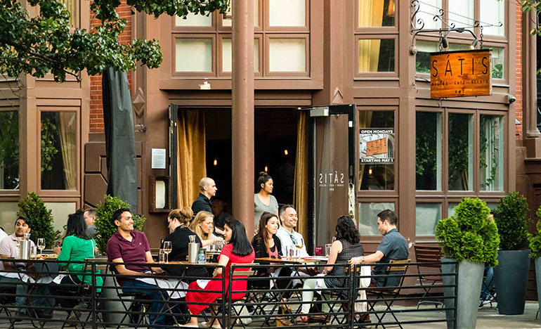 Several tables of outdoor diners enjoy Satis bistro cafe patio with laughter and wine on the table